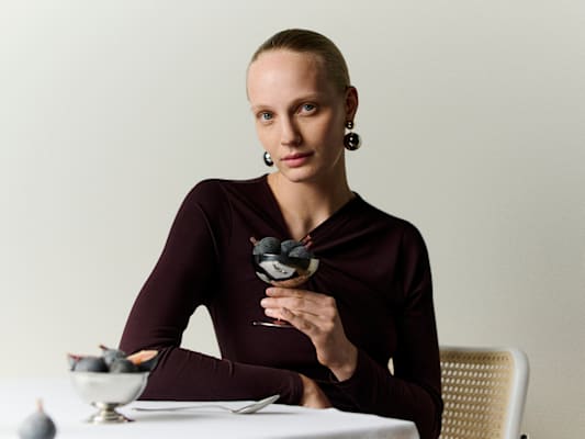 A woman in a sleeveless white outfit and sunglasses sits on a bench outdoors with shadows around her
