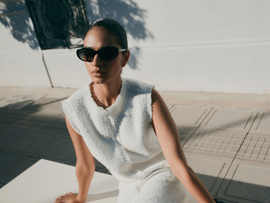 A woman in a sleeveless white outfit and sunglasses sits on a bench outdoors with shadows around her