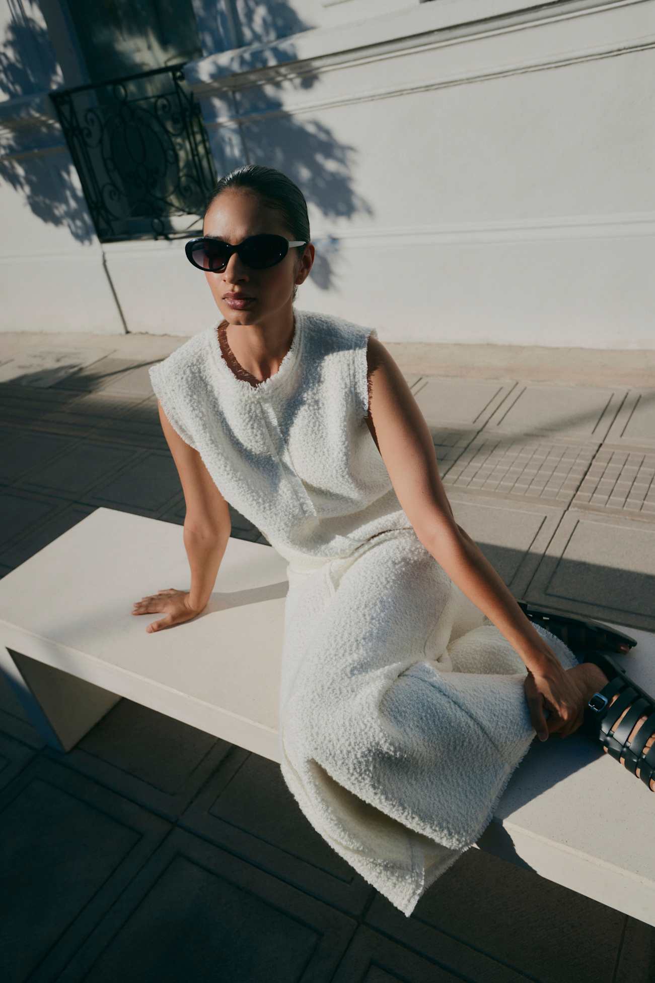 A woman in a sleeveless white outfit and sunglasses sits on a bench outdoors with shadows around her