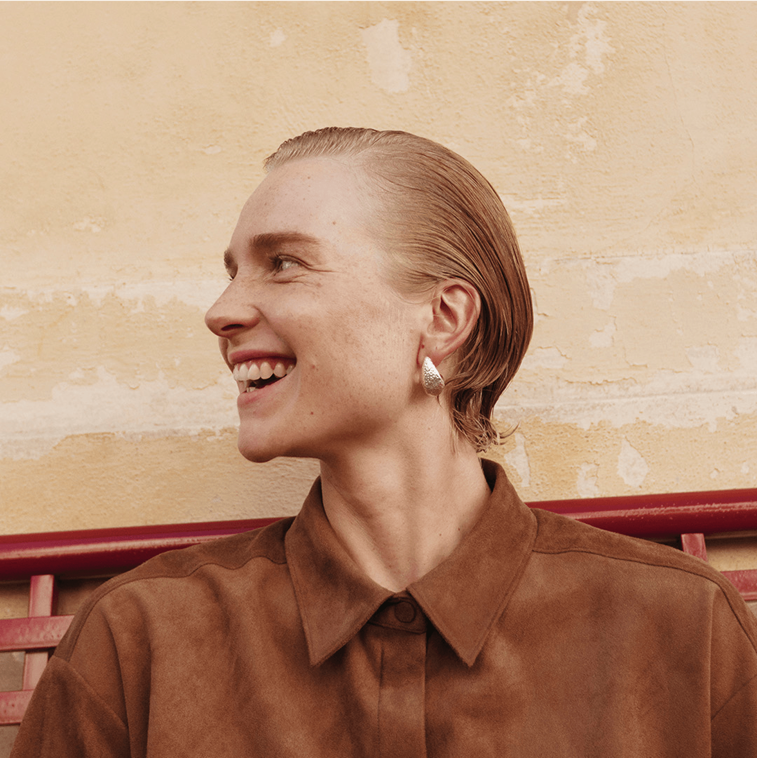 A person with short, slicked-back hair and earrings is smiling, seated in front of a light-colored wall and a red bench.