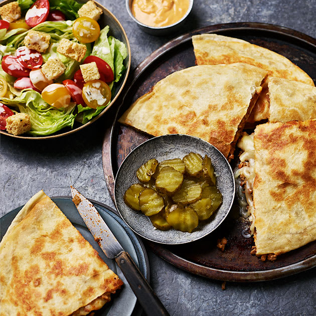 Cheeseburger quesadillas cut into quarters on a dark plate with a wedge salad and lots of pickle coins in a bowl