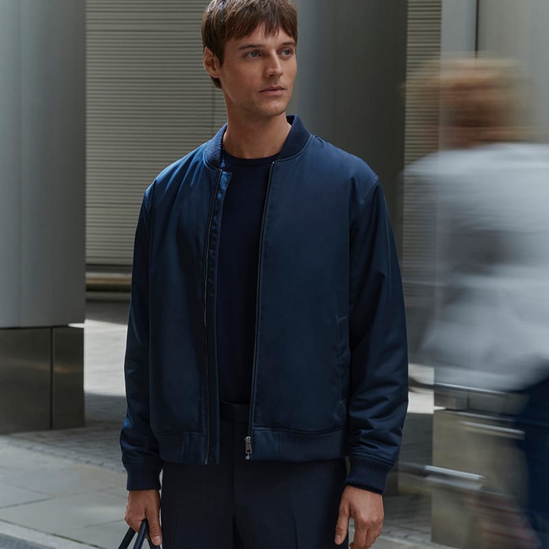 Man wearing navy regular-fit chinos, satin bomber jacket and a navy weekend bag. Shop regular-fit chinos 