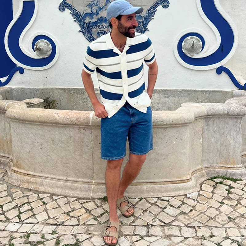 M&S Insider Dan wearing denim shorts, navy and white striped polo shirt, sandals and a baseball cap. Shop men’s shorts