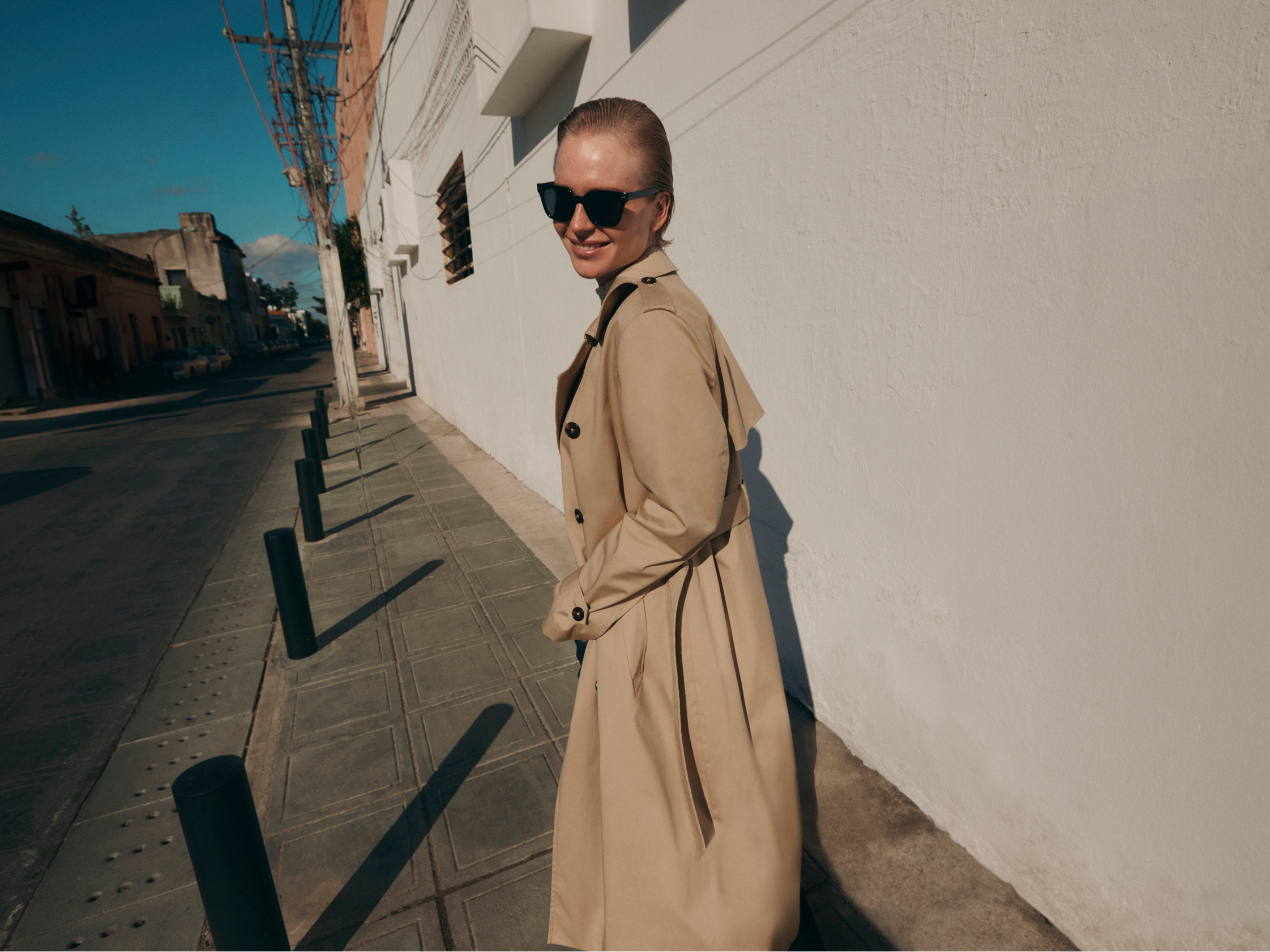A woman in a beige trench coat walking on a sunny city sidewalk, looking back at the camera with a smile.