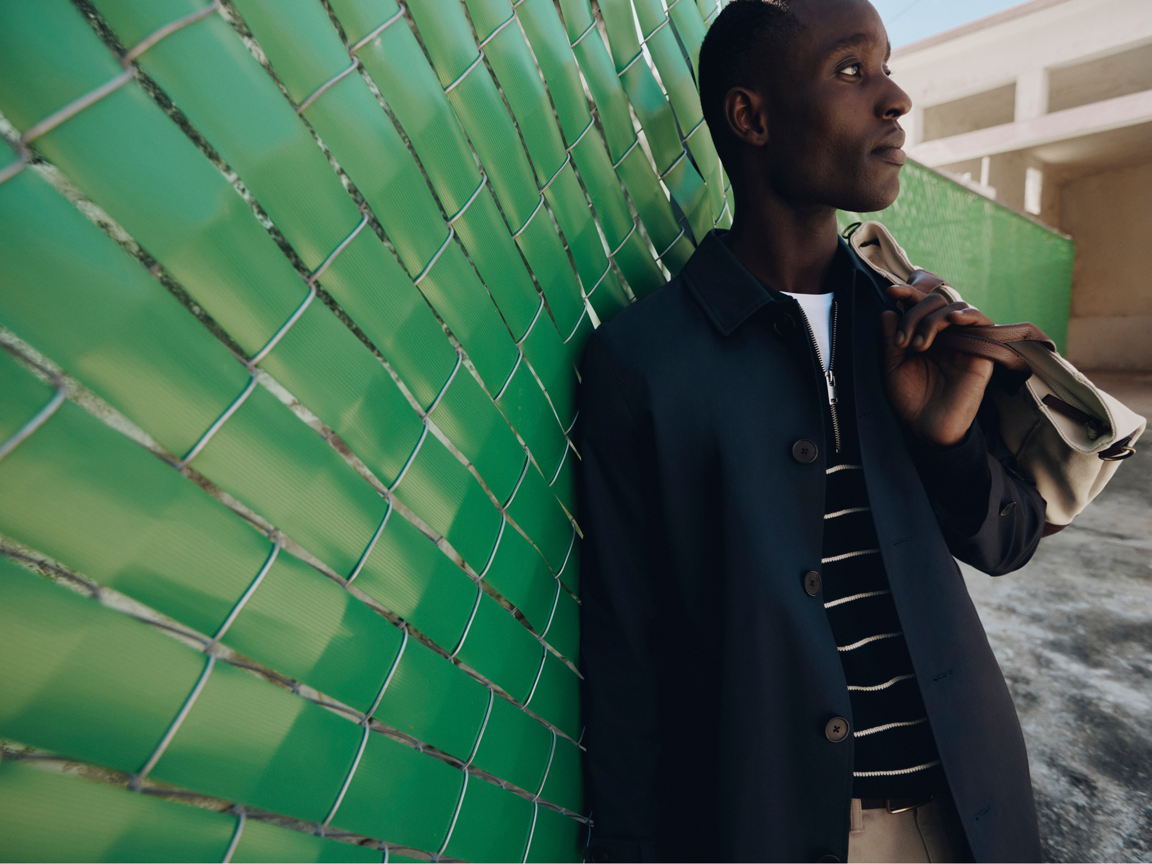 A person wearing a dark jacket and a striped shirt leans against a green wall, holding a bag over their shoulder while looking thoughtfully upwards.
