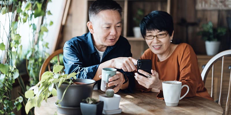 A couple shares a moment, looking at a smartphone while seated at a table with plants and mugs nearby.