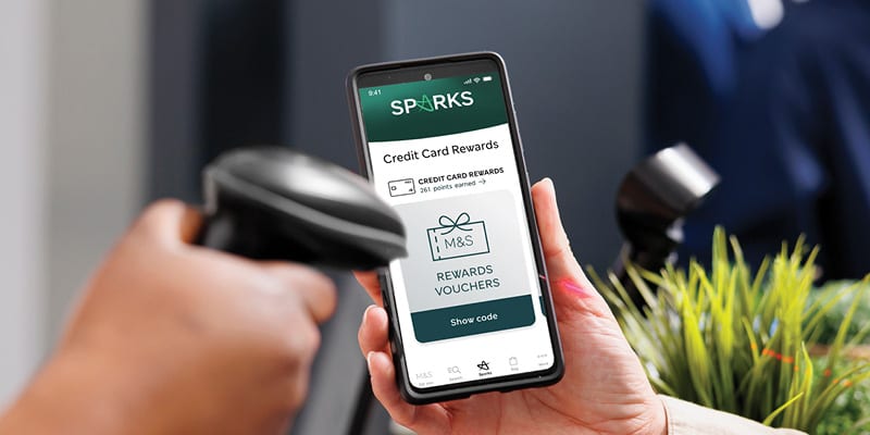 A hand holds a smartphone displaying credit card rewards, while another hand uses a barcode scanner.