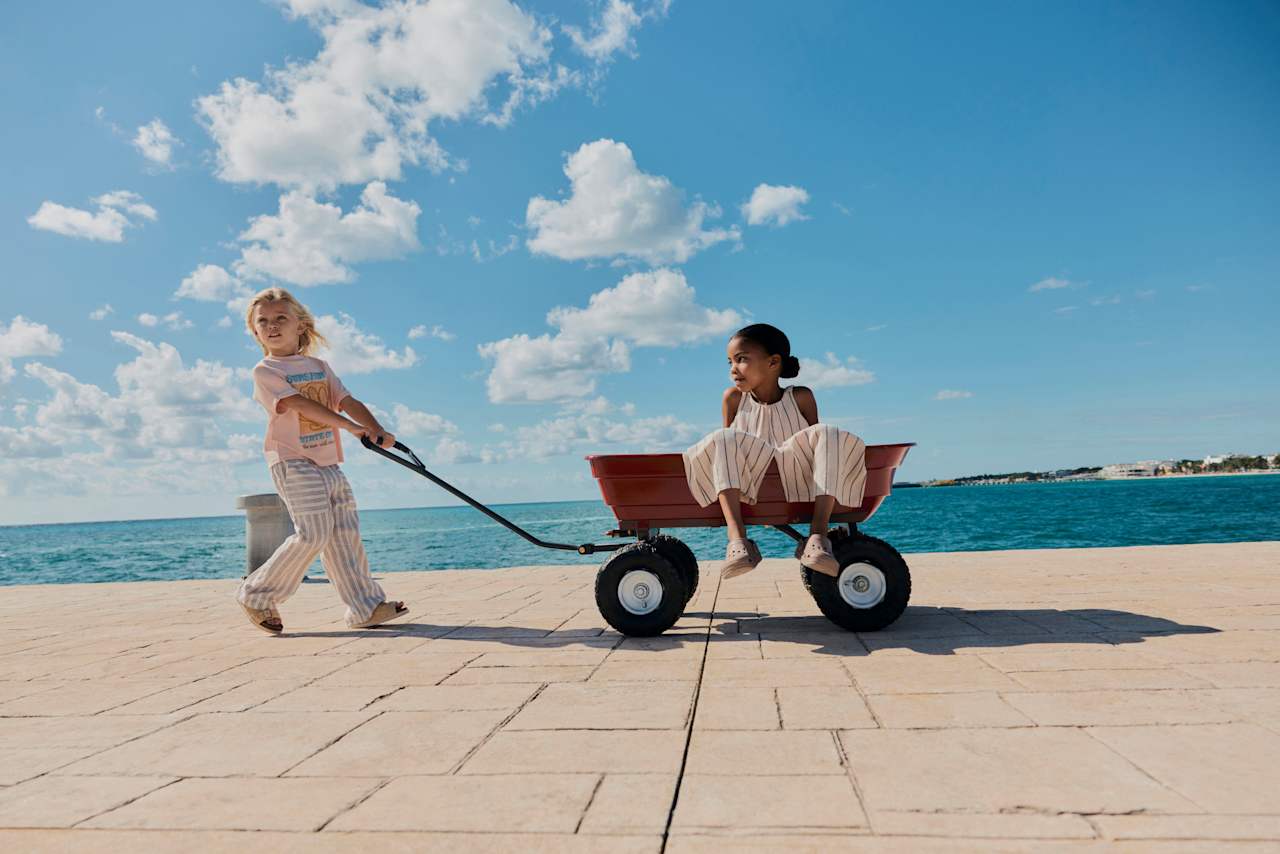 A young child with light hair pulls another young child with dark hair in a red wagon along a sunny stone promenade next to a bright blue ocean