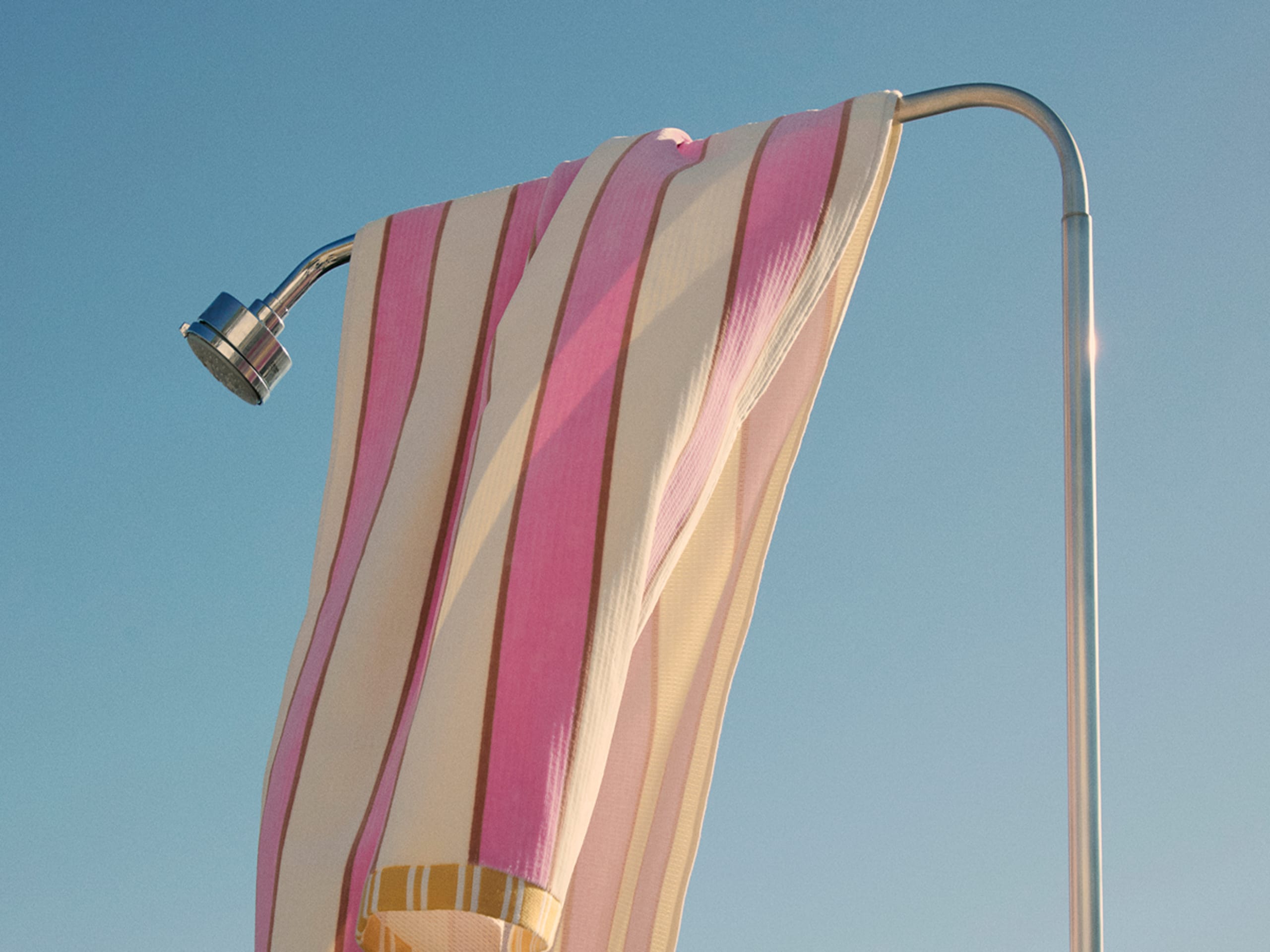 A pink and cream striped towel draped over an outdoor shower against a clear blue sky