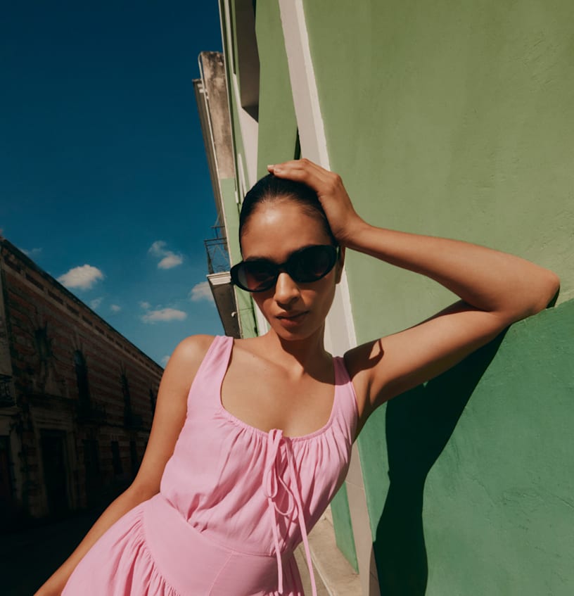 A person wearing a pink sundress with a tie-front detail and dark sunglasses is leaning against a bright lime-green building under a clear blue sky